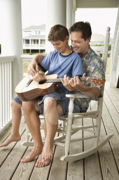 Son sits on his father's lap while playing guitar. Vertical shot.