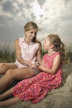 Smiling mother and daughter sit on sand looking at seashells together. Vertical shot.