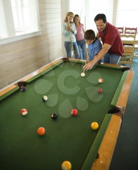 Man and boy shooting pool with woman and girl in background. Vertically framed shot.