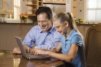 Smiling father and daughter sitting at dining room table working on laptop. 