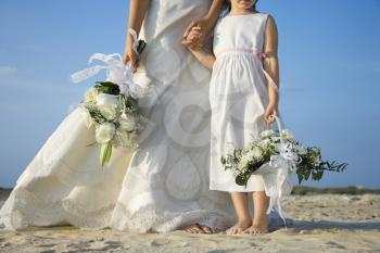 Bride and young flower girl stand on the sandy beach holding hands. Horizontal shot.