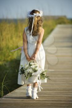 Rear view of a flower girl walking on a boardwalk holding a flower basket behind her back. Vertical shot.