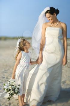 Bride and a flower girl hold hands on a sandy beach. Horizontal shot.