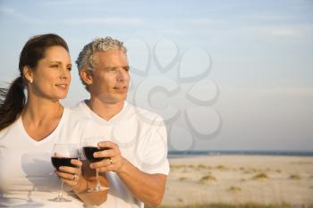 Couple drink red wine at the beach and gaze into the distance together. Horizontal shot.