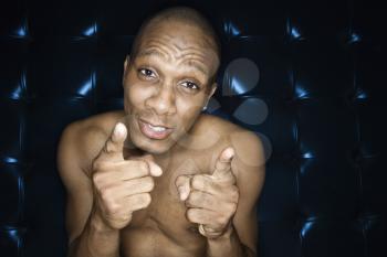 Handsome young man sitting and pointing at the camera. He is against a blue padded background, shirtless and smiling. Horizontal shot.