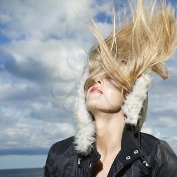 Attractive young woman is wearing a parka at the beach and flipping her long blond hair back. Square format.