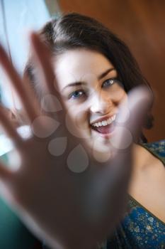Portrait of an attractive young adult woman standing by a window, holding her hand up to the camera. Vertical shot.