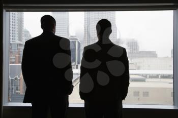 Rear view of two businessmen as they stare out a large window with a city view. They have their hands in their pockets. Horizontal view.
