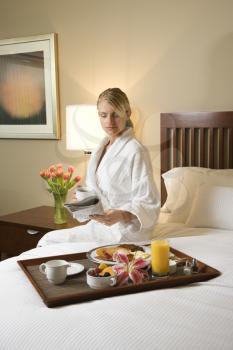 Caucasian woman wearing a bathrobe sits on a hotel bed with tray of breakfast. She is reading a newspaper and holding a coffee cup. Vertical format.