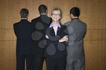 Caucasian businesswoman stands smiling as businessmen stand with their backs turned. Horizontal shot.