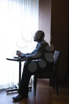 African-American businessman sitting down at a small cafe table next to a curtained window. He has a coffee cup in hand. Vertical shot.