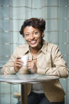 Attractive African-American woman sits at a table. She is holding a coffee cup and smiling towards the camera. Vertical shot.