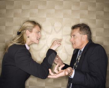 Caucasian mid-adult businesswoman yelling and pointing at middle-aged businessman, who shrugs at her. Horizontal format.