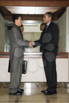 Young adult Asian and African-American businessmen standing in an office lobby shaking hands.