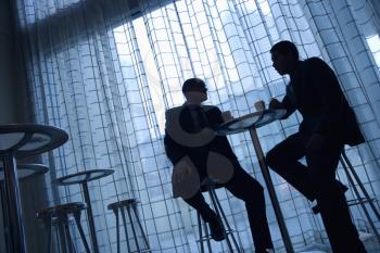 Tilt view silhouette of African-American and Asian businessmen sitting at a table having coffee in front of a curtained window.