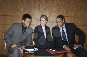 Three diverse businesspeople sit at a small table and look at a laptop together. Horizontal format.