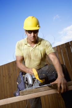Male Caucasian construction worker in safety glasses and a hardhat cutting wood with a circular saw. Vertical shot.