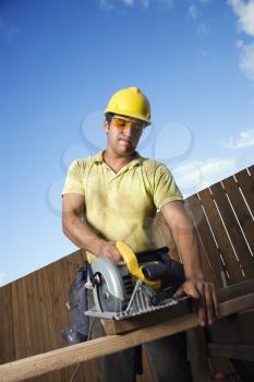 Male Caucasian construction worker in safety glasses and a hardhat. He is cutting wood with a circular saw. Vertical shot.