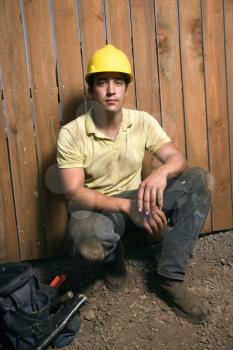 Male Caucasian construction worker squats next to a bag of tools while wearing a yellow hardhat. Vertical shot.