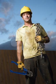 Attractive male construction worker in a yellow hardhat smiles into the camera. He holds a heavy chain on his shoulder and bolt cutters in his hand. Vertical shot.