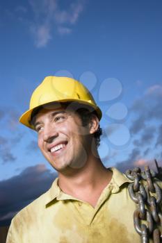 Attractive Caucasian construction worker in a yellow hardhat carries a heavy chain over his shoulder while smiling to the side. Vertical shot.