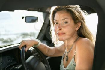 Portrait of a young Caucasian female sitting in the driver's seat of an SUV and looking at the camera, with ocean waves visible in the background. Horizontal format.
