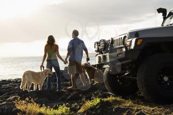 Rear view of a man and woman holding hands and relaxing with their dogs at a beach with the edge of an SUV visible in the foreground. Horizontal format.