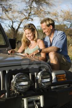 A smiling man and woman look at a map spread out on the hood of a car. Vertical format.
