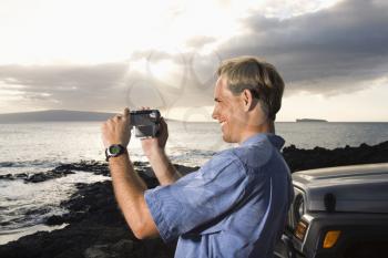 Side view of a smiling Caucasian man photographing a scenic sunset at a beach. Horizontal format.