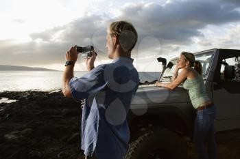 Woman leaning on an SUV while man video records scenery on a beach. Horizontal format.