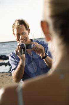 View over woman's shoulder to smiling man using video camera at the beach. Vertical format.