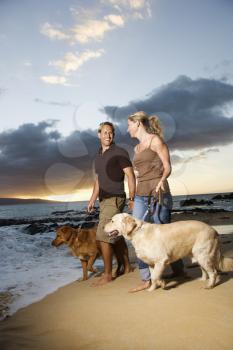 A man and woman smile at each other as they walk their dogs on a beach. Vertical format.