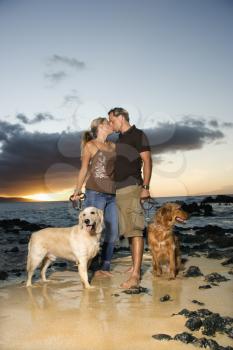 A man and woman hold the leashes of their dogs as they kiss on a beach. Vertical format.