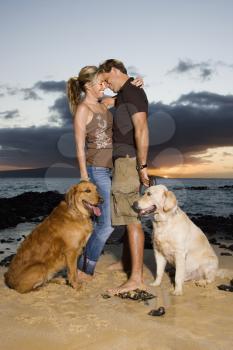 A man and woman hold the leashes of their dogs as they hug at a beach. Vertical format.
