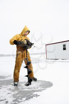 Young man in yellow snow gear smiles as he drills a hole in the ice for fishing. Vertical shot.