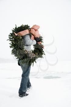 Adult male walks through the snow in a winter landscape while carrying a decorated wreath. Vertical shot