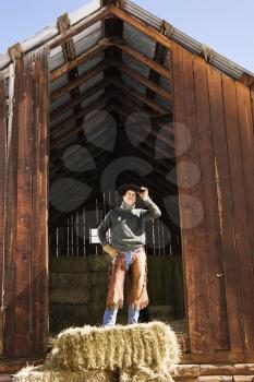 Attractive young man wearing a cowboy hat while standing on top of hay bales. He is tipping up his hat. Vertical shot.