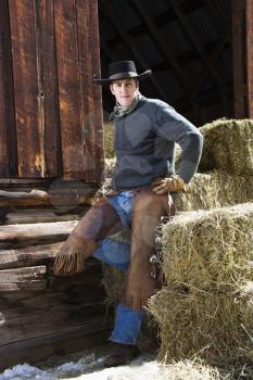 Attractive young man wearing a cowboy hat and chaps. He is leaning on hay bales outside of a barn. Vertical shot.
