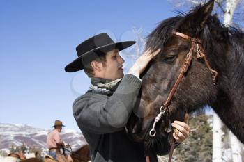 Attractive young man wearing a cowboy hat. He is petting a horse with another rider in the background. Horizontal shot.