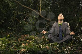 Young businessman sits in a lotus position meditating in the woods with closed eyes and a smile. Horizontal shot.