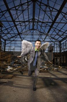 Young businessman smiles with angel wings on his back and attempts takeoff in an abandoned building. Vertical shot.