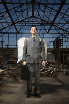 Young businessman with angel wings smiles at the camera while standing in an abandoned building. Vertical shot.