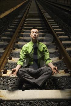Young businessman sits in a lotus position and meditates while on railroad tracks. Vertical shot.