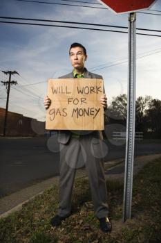 Young businessman stands on a street corner holding a sign that reads 'will work for gas money'. Vertical shot.