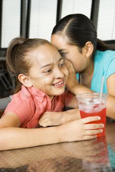 Asian girl whispers into the ear of a smiling Hispanic girl with a beverage. Vertical shot.
