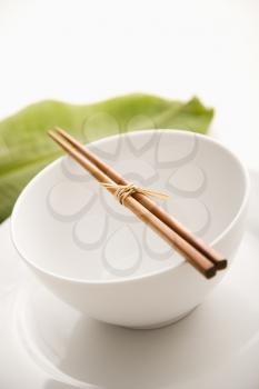 Chopsticks lying across an empty bowl on top of a plate. The dishes are white, and there is a green leaf in the background. Vertical shot. Isolated on white.