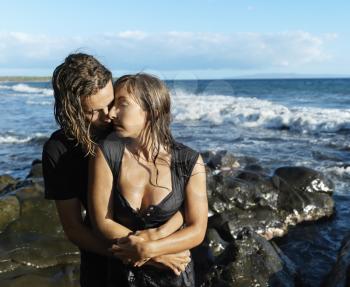Attractive young man embraces a woman from behind while standing on a rocky beach. Horizontal shot.
