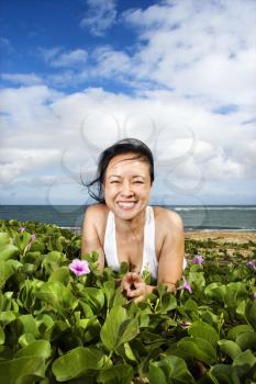 Smiling asian woman in a patch of flowers and foliage with the beach and ocean in the distance. Vertical shot.