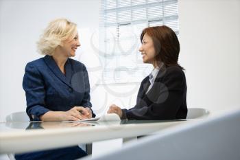 Two businesswomen sitting at office desk looking at eachother smiling.
