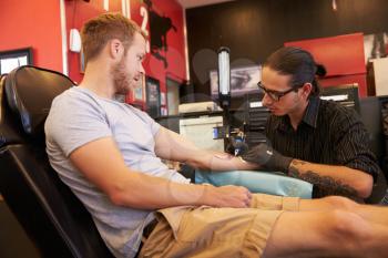 Man Sitting In Chair Having Tattoo On Arm In Parlor